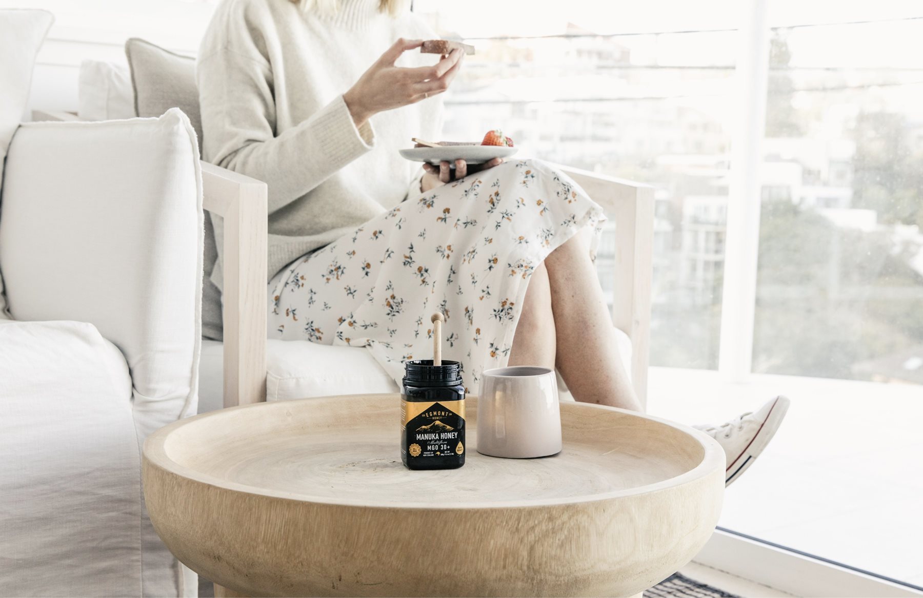 A slightly out of focus lady eating toast in a white dress.  The chair is also white.  In the foreground is a round concave wooden table showing a jar of Mānuka Honey 