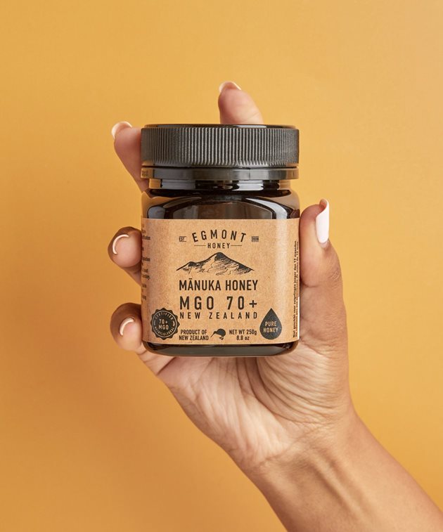 A hand holding a bottle of Mānuka Honey.  The bottle is dark brown glass with a light brown label.  On the label is a black illustration of a mountain, reflecting the company's New Zealand location