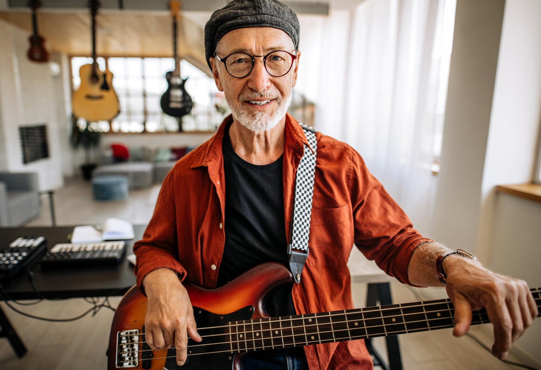 A Senior musican with a flat cap and short white hair and glasses sits holding his guitar from a home studio