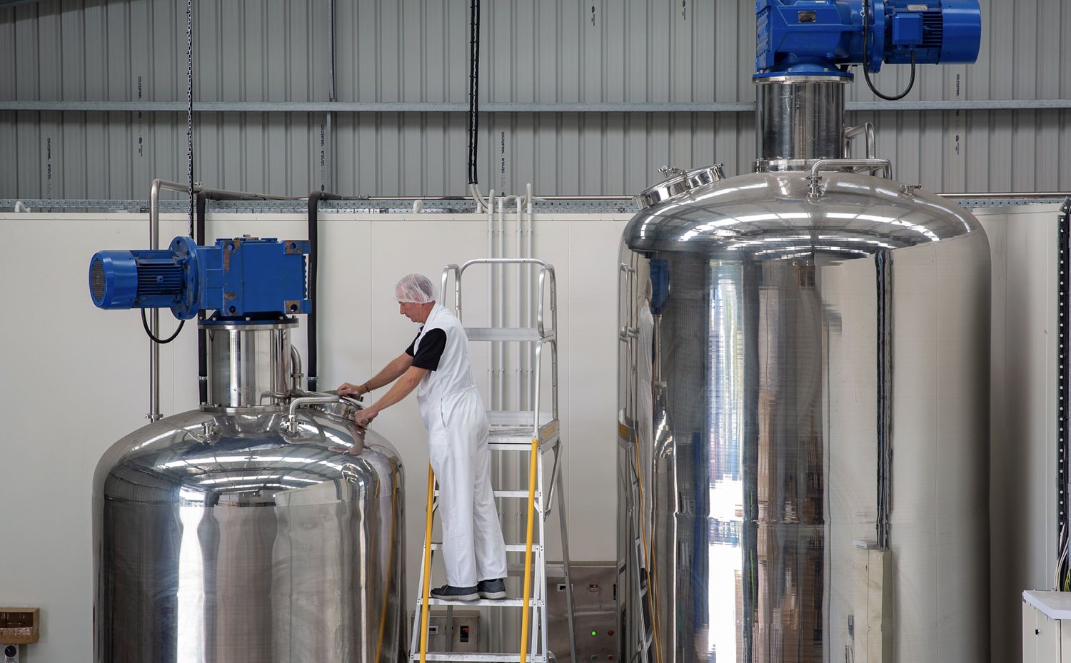 A shot of a man in a warehouse.  He's up a ladder next to two giant chrome vats of honey.  He appears to be about to open one of them