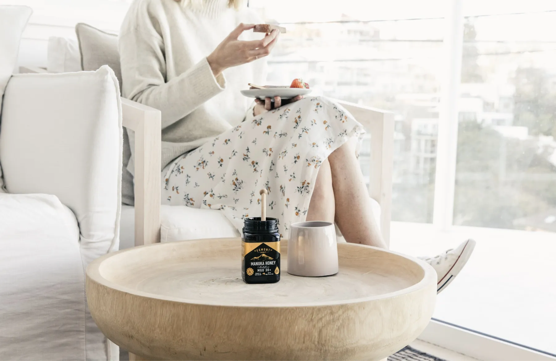 A slightly out of focus lady eating toast in a white dress. The chair is also white. In the foreground is a round concave wooden table showing a jar of Mānuka Honey